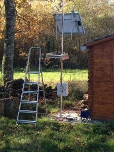 Station météo sur un mât, avec son panneau solaire, à côté d'une cabane en bois.