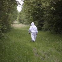 Apiculteur en combinaison blanche marchant sur un chemin herbeux entre les arbres.